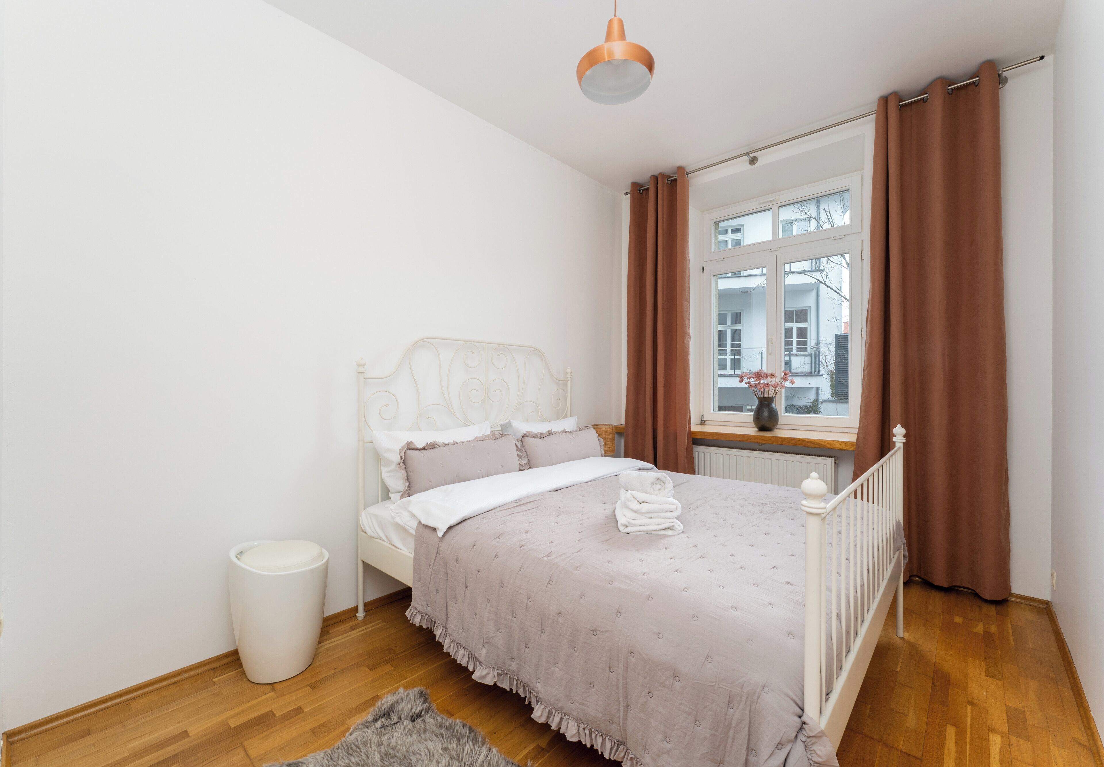 A bedroom with a bed, a window with curtains, a radiator, and a trash bin.