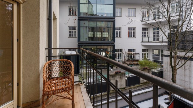 Un balcone con una sedia in vimini, vista su un edificio moderno con ampie finestre e ringhiera metallica.