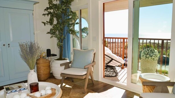 A room with a view of the beach, a chair, a table with decorative items, and a large plant. - Oceanfront Sanctuary in Hilton Head Island (Hilton Head Island)