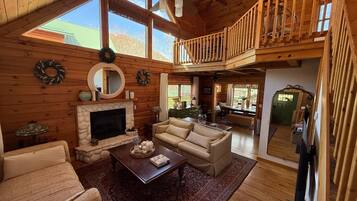 A spacious living room with a fireplace, a high ceiling with a balcony, and a view of the outside.
