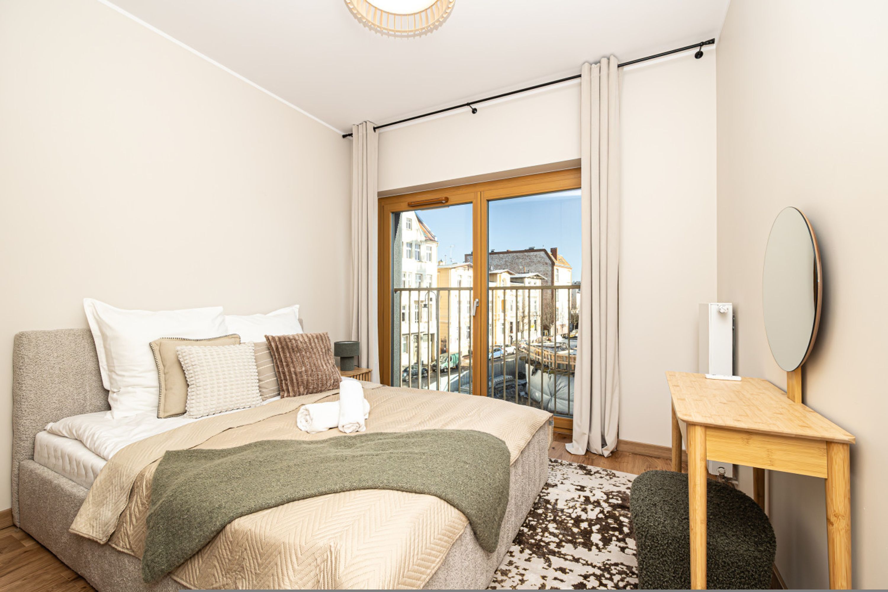 A hotel room with a bed, a bedside table, a mirror, and a view of buildings through a large window.