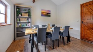 A dining area with a wooden table set for four, surrounded by chairs, a bookshelf with plants, and a painting on the wall. - Waterside Cottage in Pancrasweek (Holsworthy)