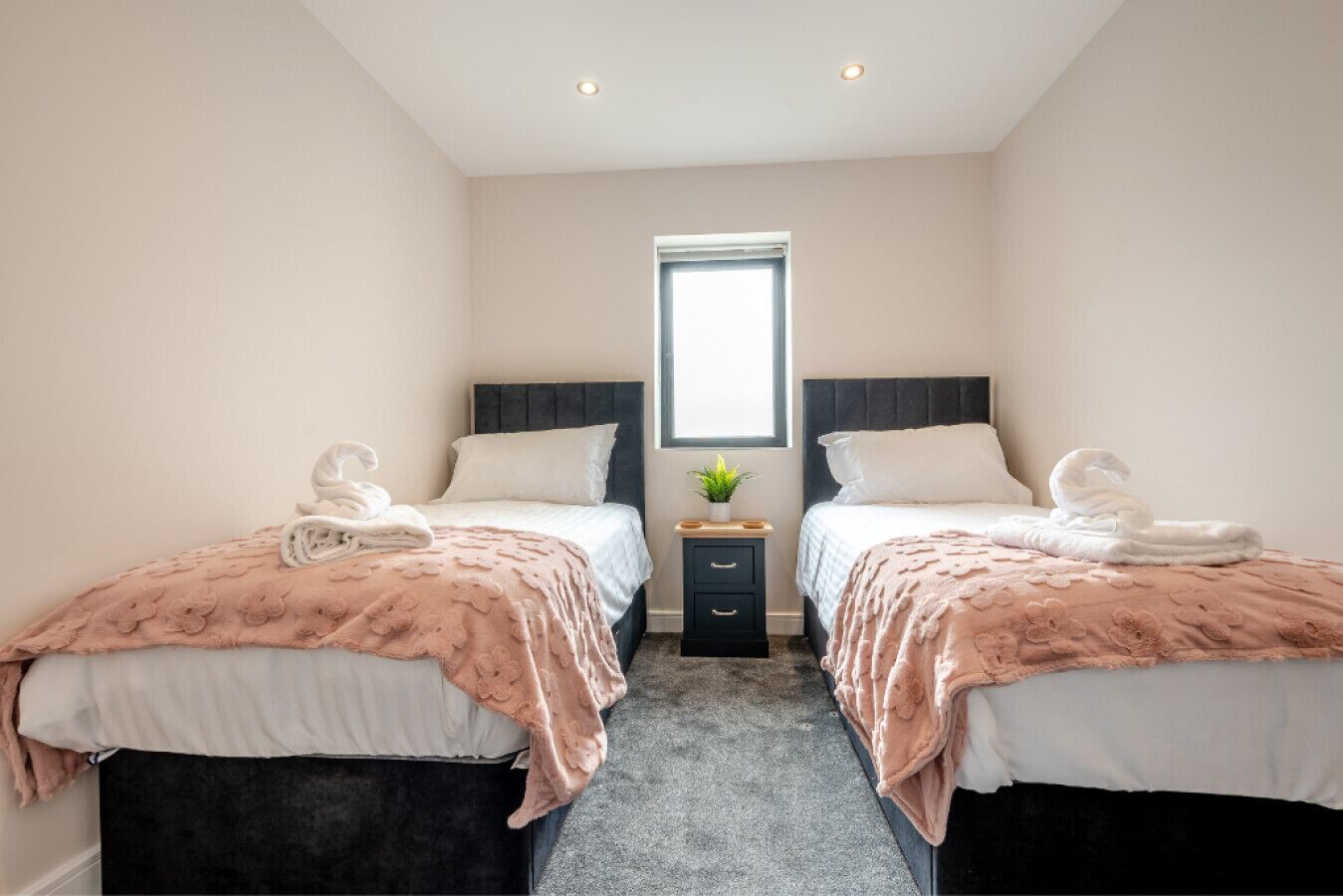 A hotel room with two beds, each with a pink floral bedspread and white pillows, a small bedside table with a plant, and a window with a view.
