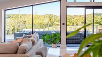 Una sala de estar moderna con un sofá grande, una mesa de centro y una planta en maceta. Hay una puerta corrediza de vidrio que da a un balcón con mobiliario exterior y vistas a un área boscosa.
