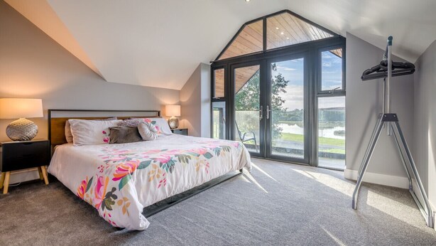 Une chambre à coucher avec un grand lit, une table de chevet, une lampe et un trépied. Une grande fenêtre offre une vue sur un lac et des arbres.
