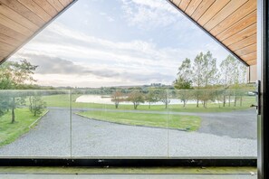 A view through a triangular window of a gravel path leading to a lake, surrounded by trees and a grassy area. - Lake View House Pancrasweek (Holsworthy)