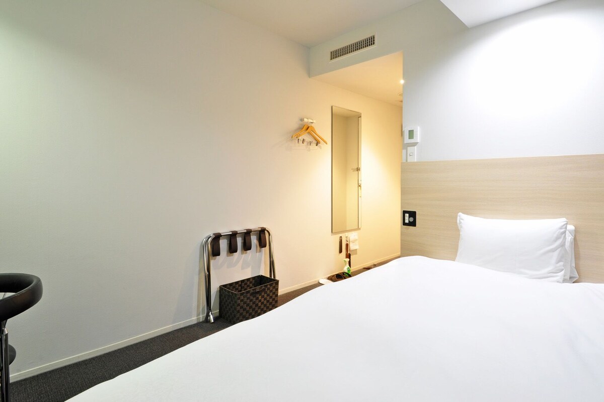 Ein Hotelzimmer mit Bett, Sessel, Kleiderhaken, Spiegel und einem kleinen Tisch mit einer Flasche.
