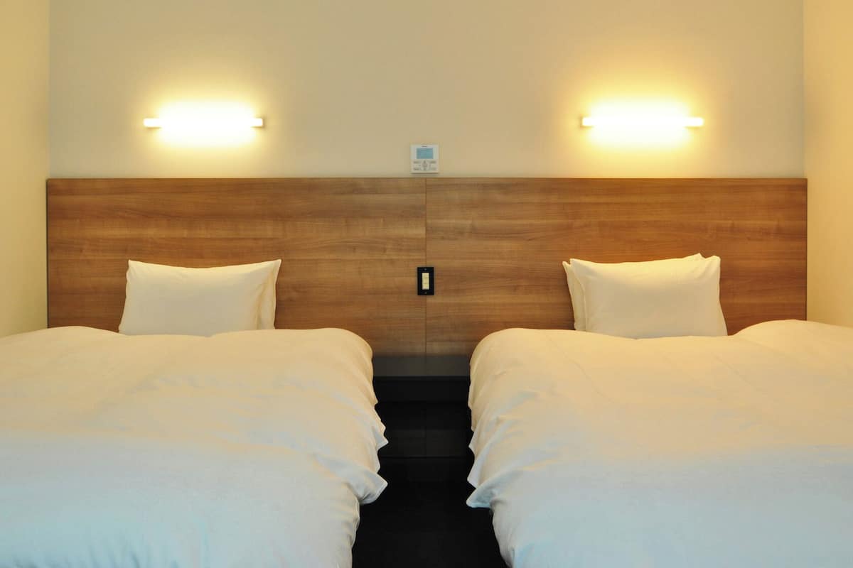 A hotel room with two beds, wooden headboard, wall-mounted lights, and a small nightstand.