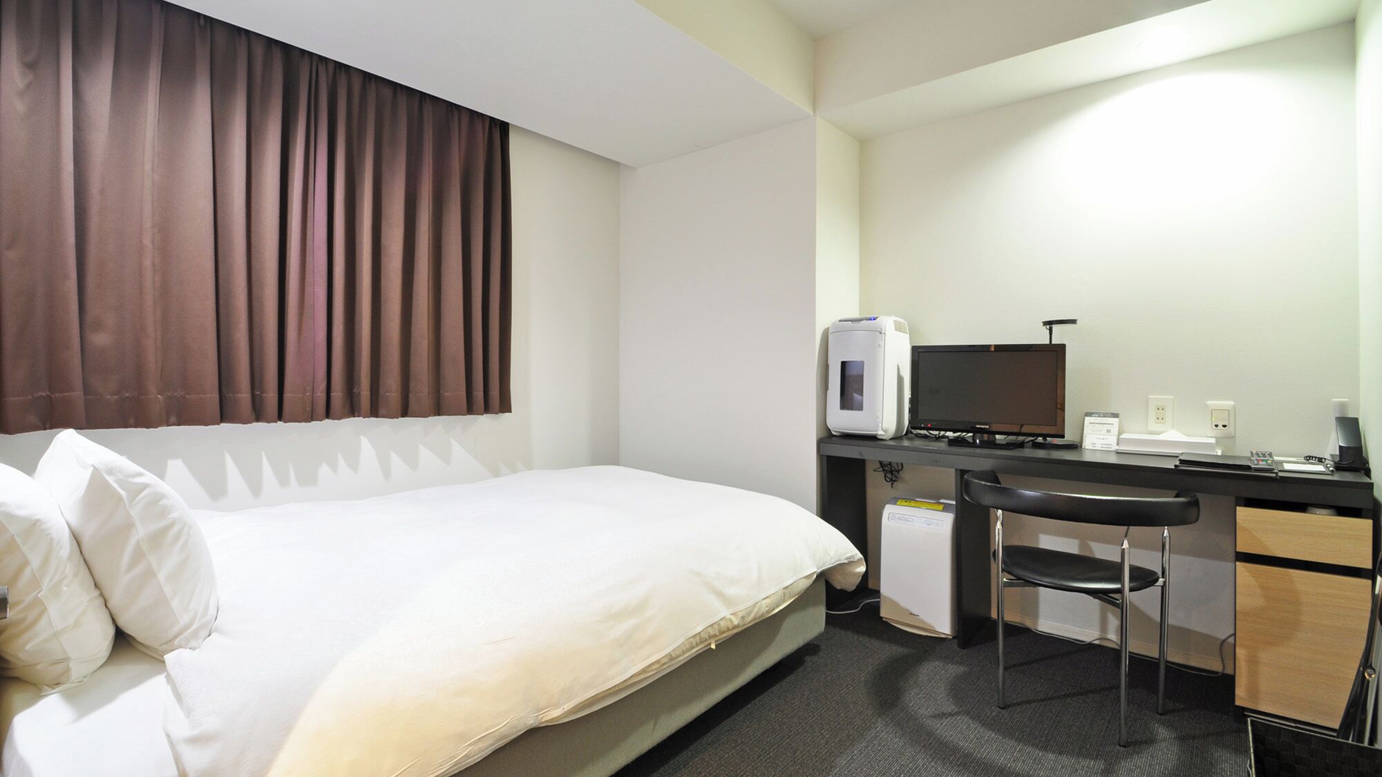 Ein Hotelzimmer mit Bett, Schreibtisch mit Computer, Stuhl und einem Fenster mit Vorhängen.