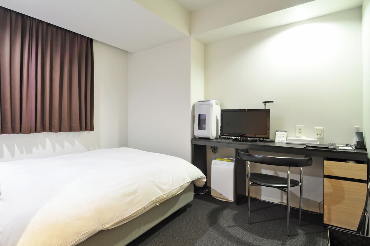 A hotel room with a bed, a desk with a computer, a chair, and a window with curtains.