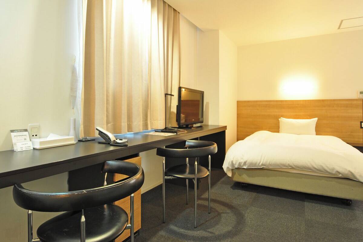 A hotel room with a bed, a desk with a computer, two bar stools, a television, and a window with curtains.
