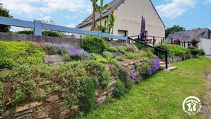 Una casa con techo inclinado, rodeada de un jardín con diversas plantas y flores.