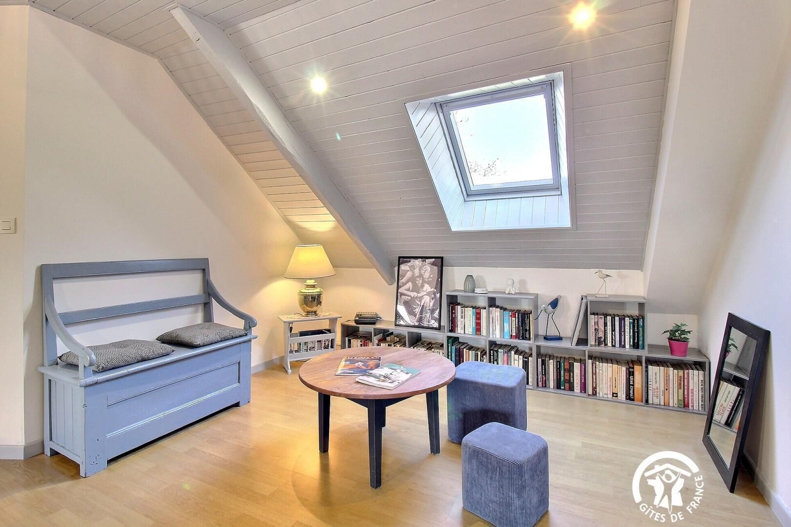 Una habitación estilo loft acogedora, con techo inclinado, tragaluz, sofá azul, una mesa redonda y una estantería llena de libros.
