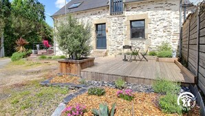 Una casa de piedra con terraza de madera, mobiliario exterior y un jardín con flores y plantas.
