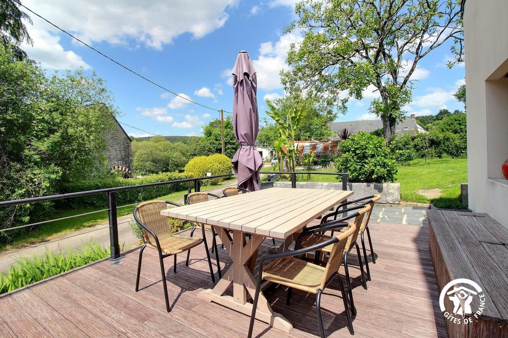 An outdoor patio with a wooden table and wicker chairs, surrounded by greenery and a clear sky.