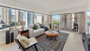 Una sala moderna con sofá, reposapiés y mesa de centro, ventanas amplias con vistas a la ciudad y un balcón con vista a edificios de gran altura.