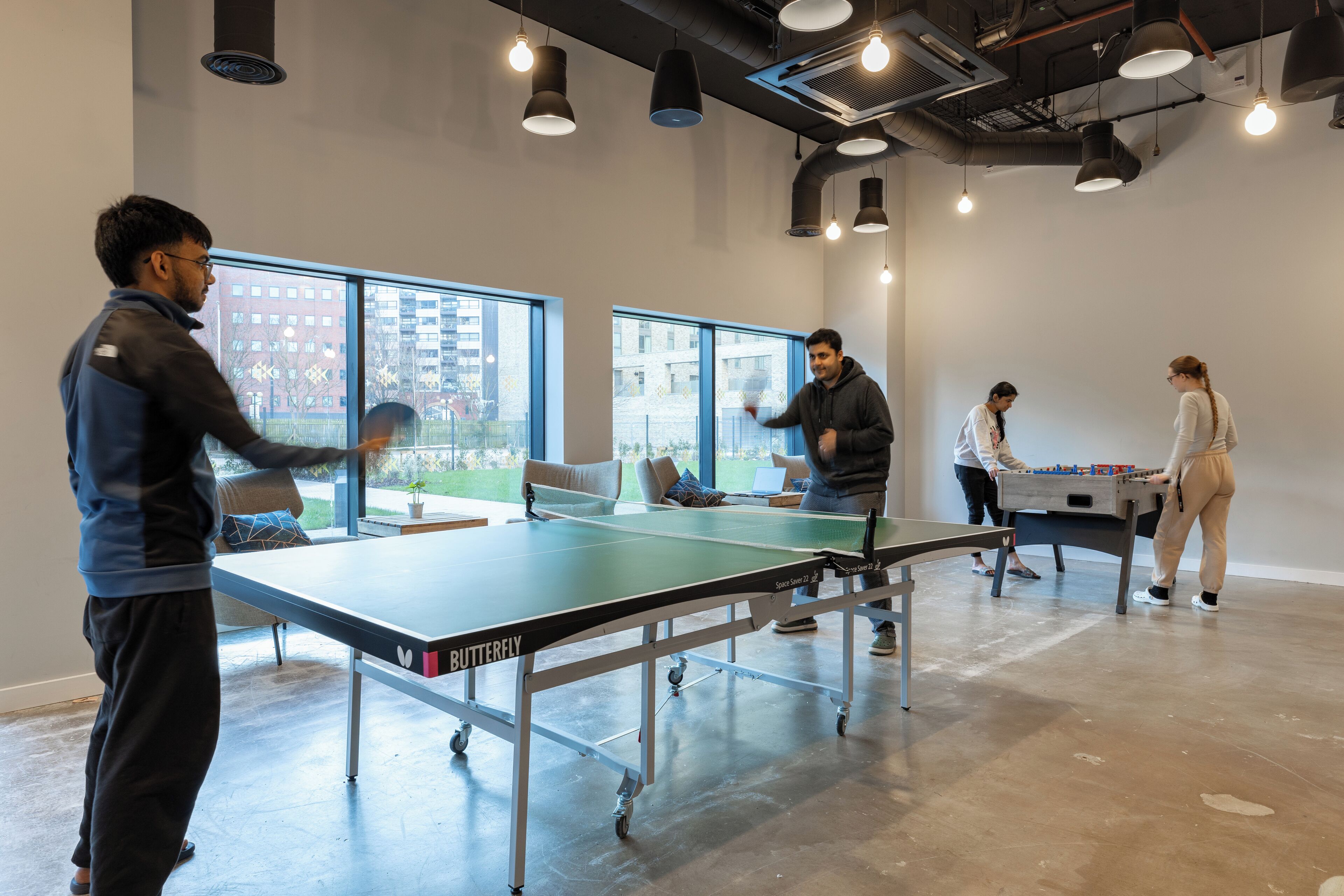 A modern indoor space with a ping pong table, a foosball table, and large windows offering views of buildings outside.