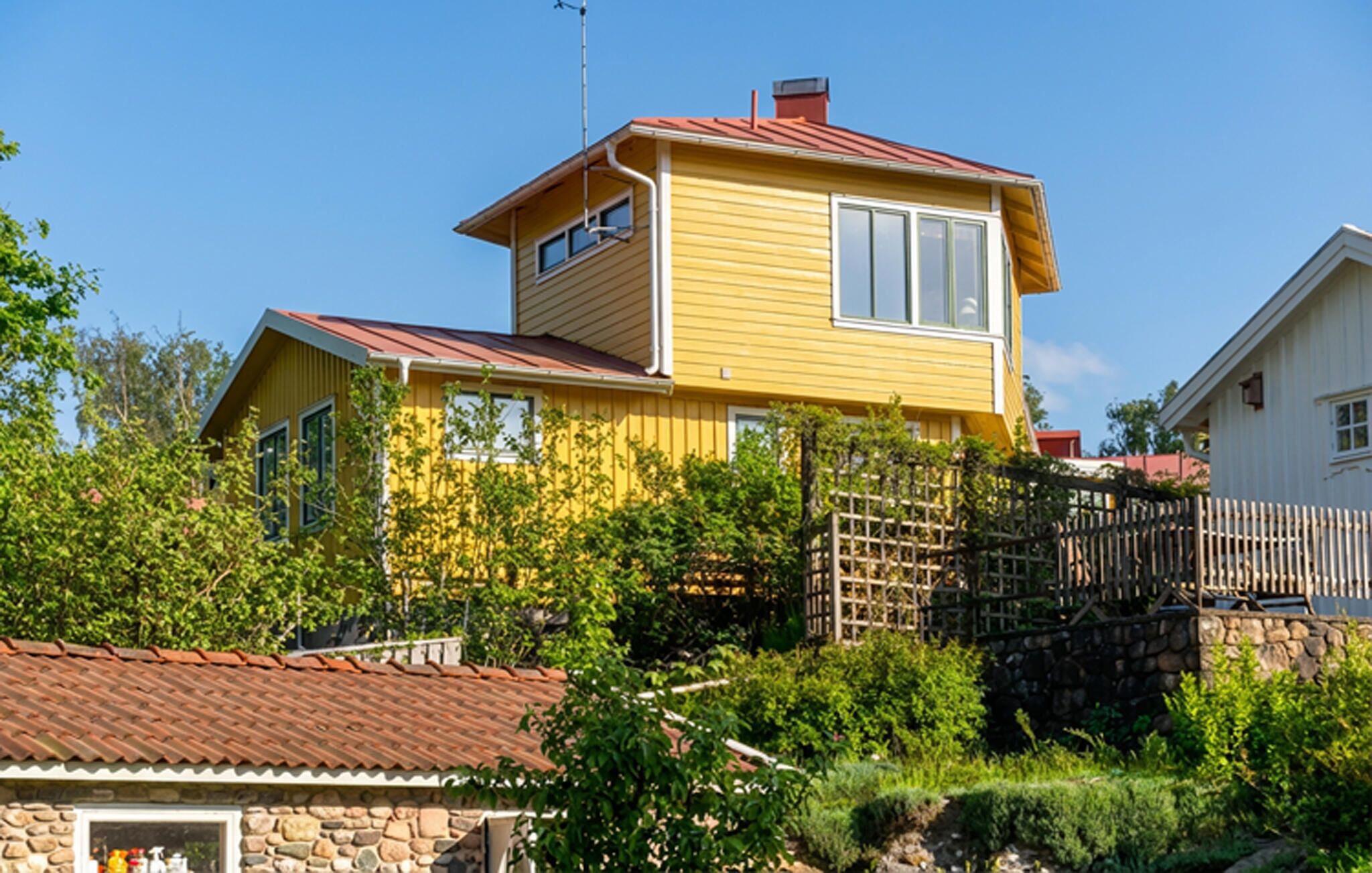 A two-story yellow house with a red roof, surrounded by greenery and a stone wall.