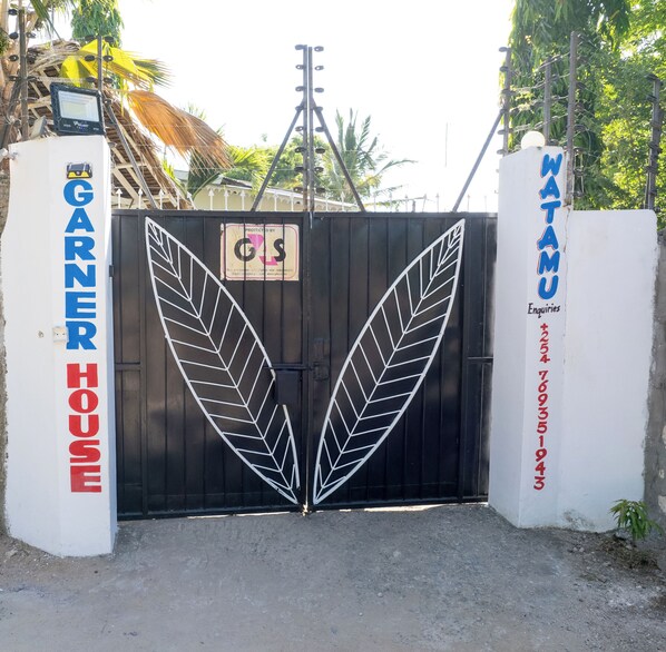 A gated entrance with a large leaf-like design, a sign indicating 'GARNER HOUSE' on the left, and 'GATAMU Enquiries' on the right.