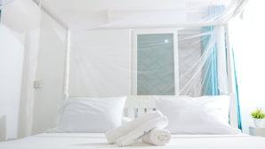 Ein ordentlich bezogenes Bett mit weißen Leinen und Kissen, ein weißes Moskitonetz und ein Fenster mit blauen Vorhängen.