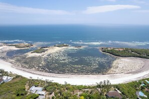 Vue aérienne d’une zone côtière avec une plage, des formations rocheuses et quelques bâtiments dispersés.