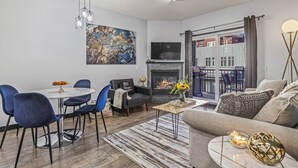 A modern living room with a fireplace, a dining area with blue chairs, and a large window with a view of the outside. - Perfect Rockies Escape with Pool, Hot Tub and Gym (Canmore)