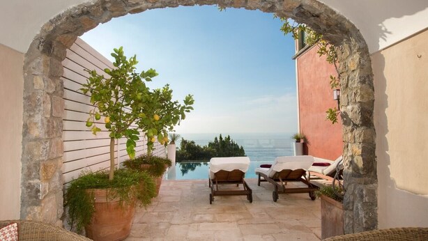 Un patio al aire libre con un arco de piedra, dos sillones de descanso, una piscina pequeña y vista al mar.
