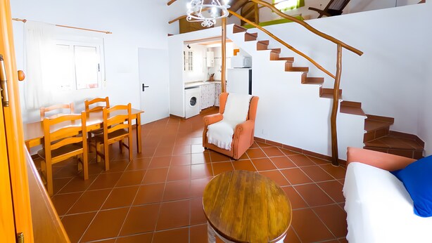 Amplia sala de estar con una mesa de comedor, sillas, un sofá, una escalera de madera y una cocina al fondo.