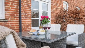 Un espace de restauration en plein air avec des chaises en osier, une table en verre, une miche de pain, un bol de chips et un vase de fleurs.