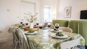 Un coin repas avec une table dressée pour quatre personnes, entourée de chaises, d’un vase de fleurs et d’une fenêtre laissant entrer la lumière naturelle.