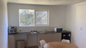 A compact kitchen with a microwave, sink, and small trash bin. There is a round wooden table with four chairs and a window above it. - Country cottage in the heart of the Yarra Valley (Seville)