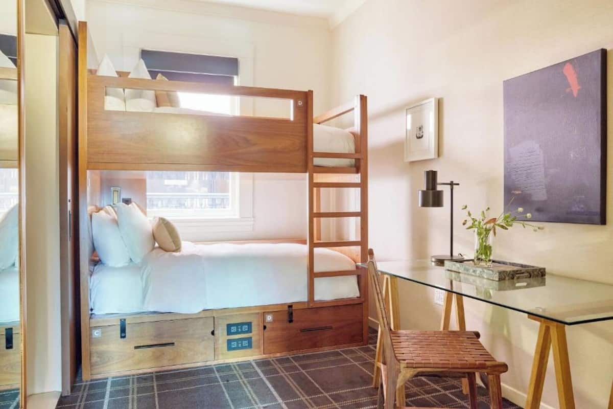 A bunk bed room with a desk and chair, a window, and a painting on the wall.