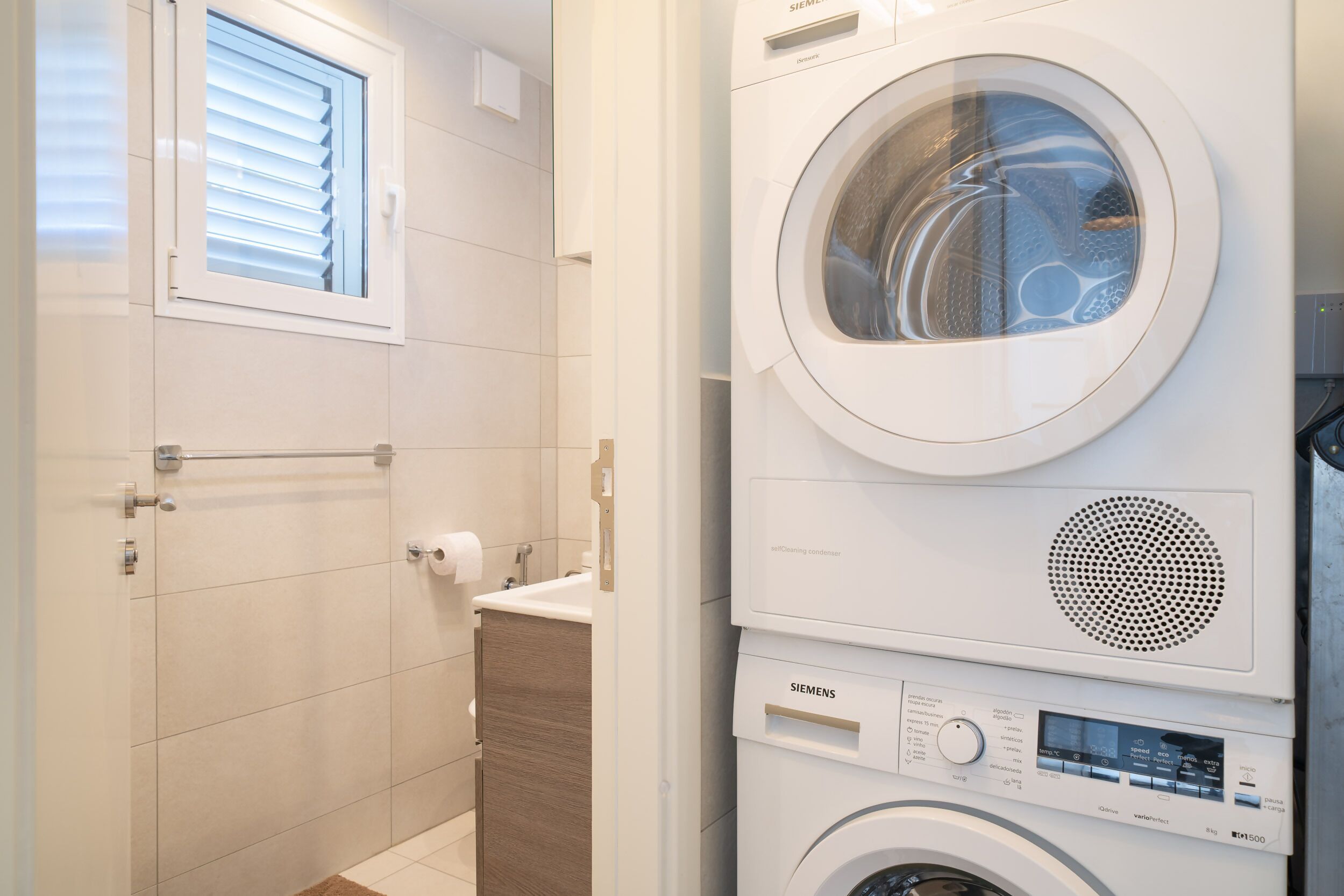 Une buanderie moderne avec une machine à laver et un sèche-linge superposés, une fenêtre avec des stores et un meuble en bois.