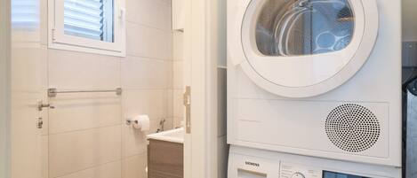 Une buanderie moderne avec une machine à laver et un sèche-linge superposés, une fenêtre avec des stores et un meuble en bois.