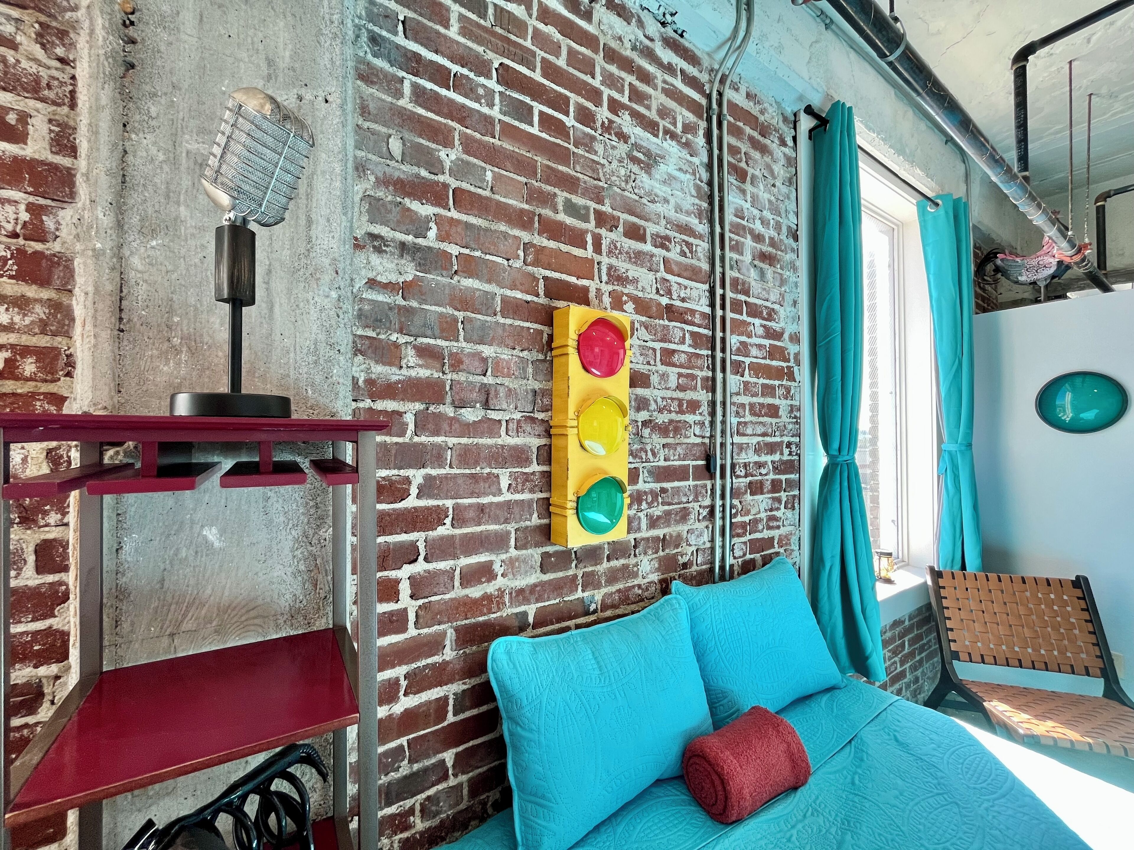 Ein Zimmer mit Backsteinwand, einem roten Regal, einem blauen Sofa mit rotem Kissen und einer Ampel-Dekoration.