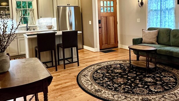 A spacious living room with a round rug, a dining area with a wooden table and chairs, a kitchen with white cabinets and a refrigerator, and a chandelier.