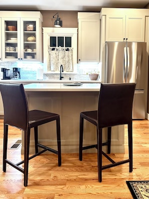 Une cuisine avec deux tabourets de bar en bois foncé, un réfrigérateur en acier inoxydable et une fenêtre avec un rideau.