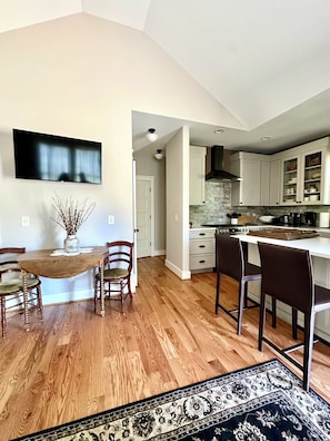 A modern kitchen with white cabinets, a dining area with a wooden table and chairs, and a large flat-screen TV mounted on the wall.