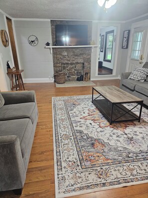 A living room with a stone fireplace, grey sofas, a patterned rug, and a television mounted above the fireplace.