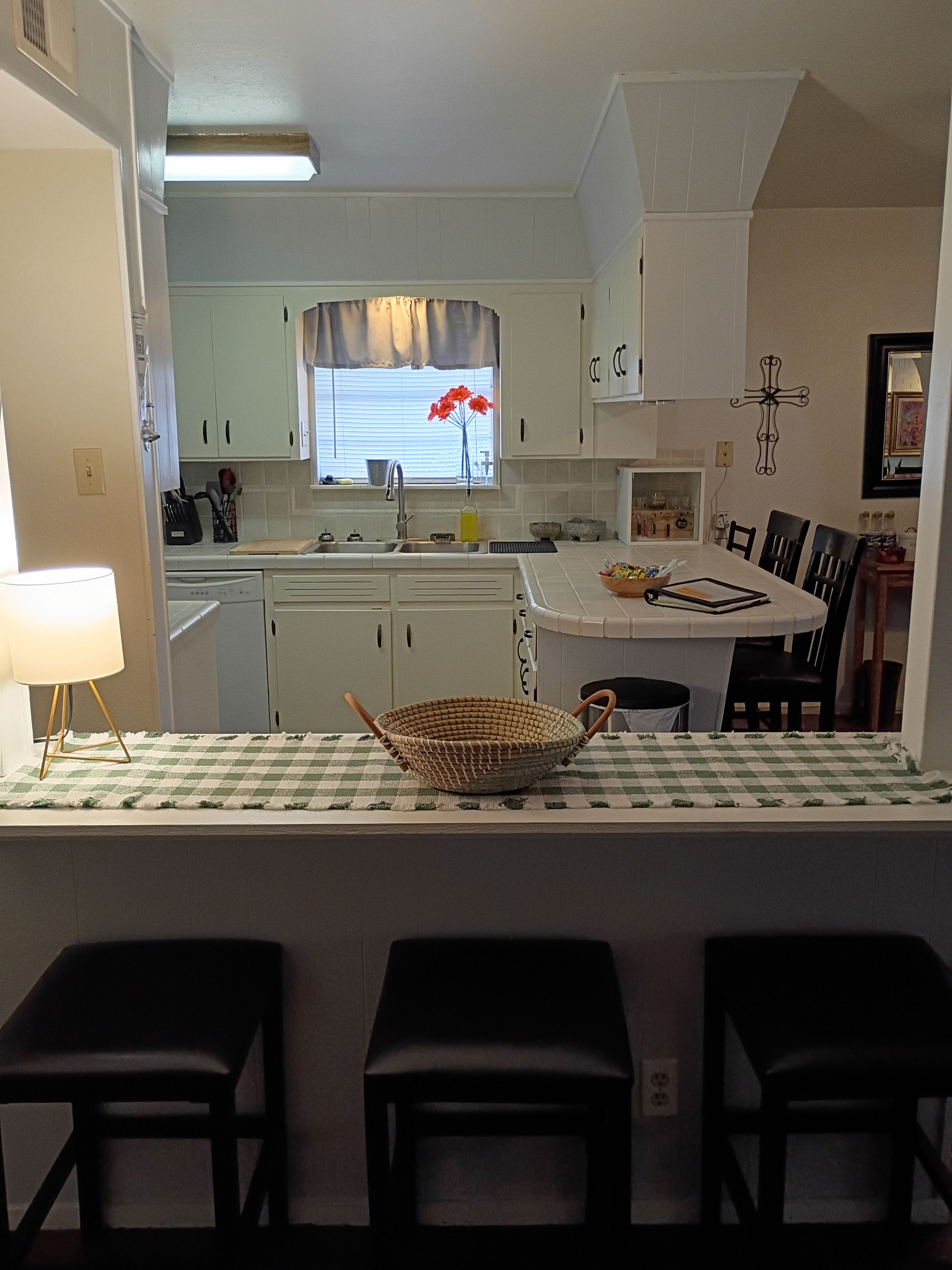 Une cuisine avec des meubles blancs, une fenêtre avec des rideaux, un évier et un coin repas avec une nappe à carreaux et des chaises.