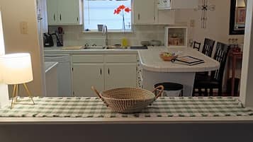 Une cuisine avec des meubles blancs, une fenêtre avec des rideaux, un évier et un coin repas avec une nappe à carreaux et des chaises.