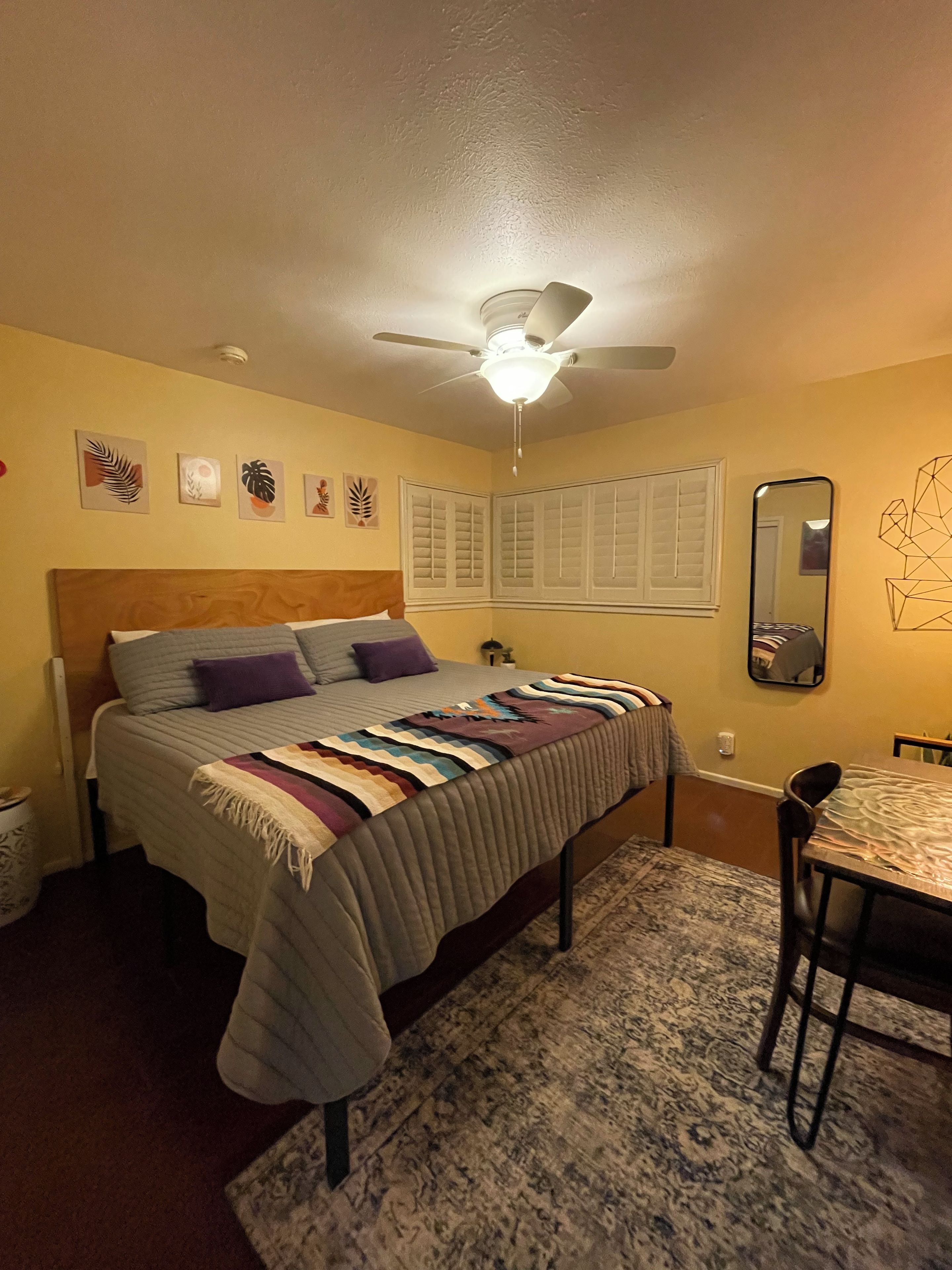 Een slaapkamer met een bed, een plafondventilator, een raam met luiken en een spiegel aan de muur.