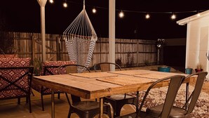Um pátio ao ar livre com mesa de madeira, cadeiras de metal, uma rede pendurada e luzes de Natal.