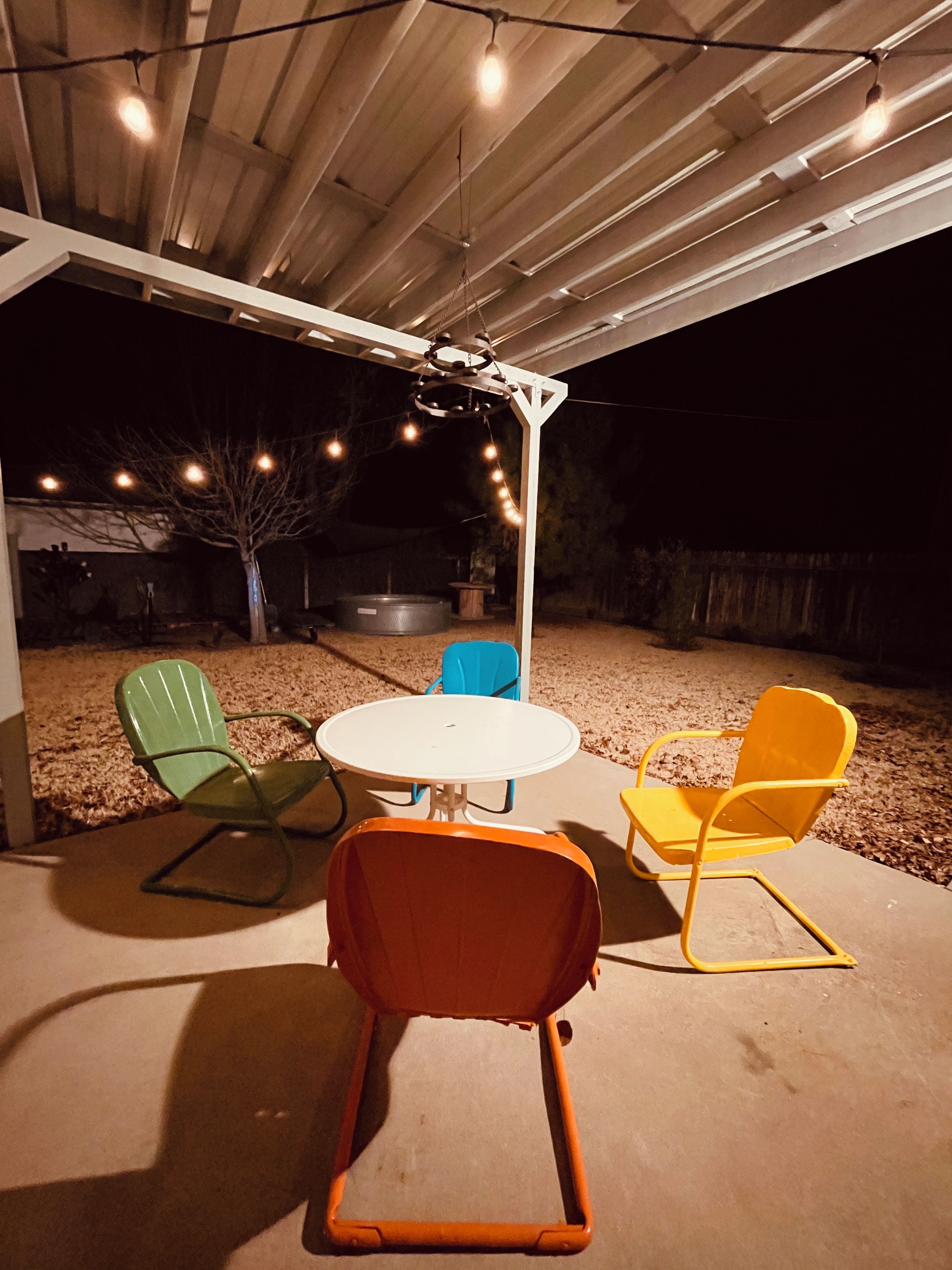 Een terras buiten met een ronde tafel en vier stoelen, een hangende lamp en een boom.