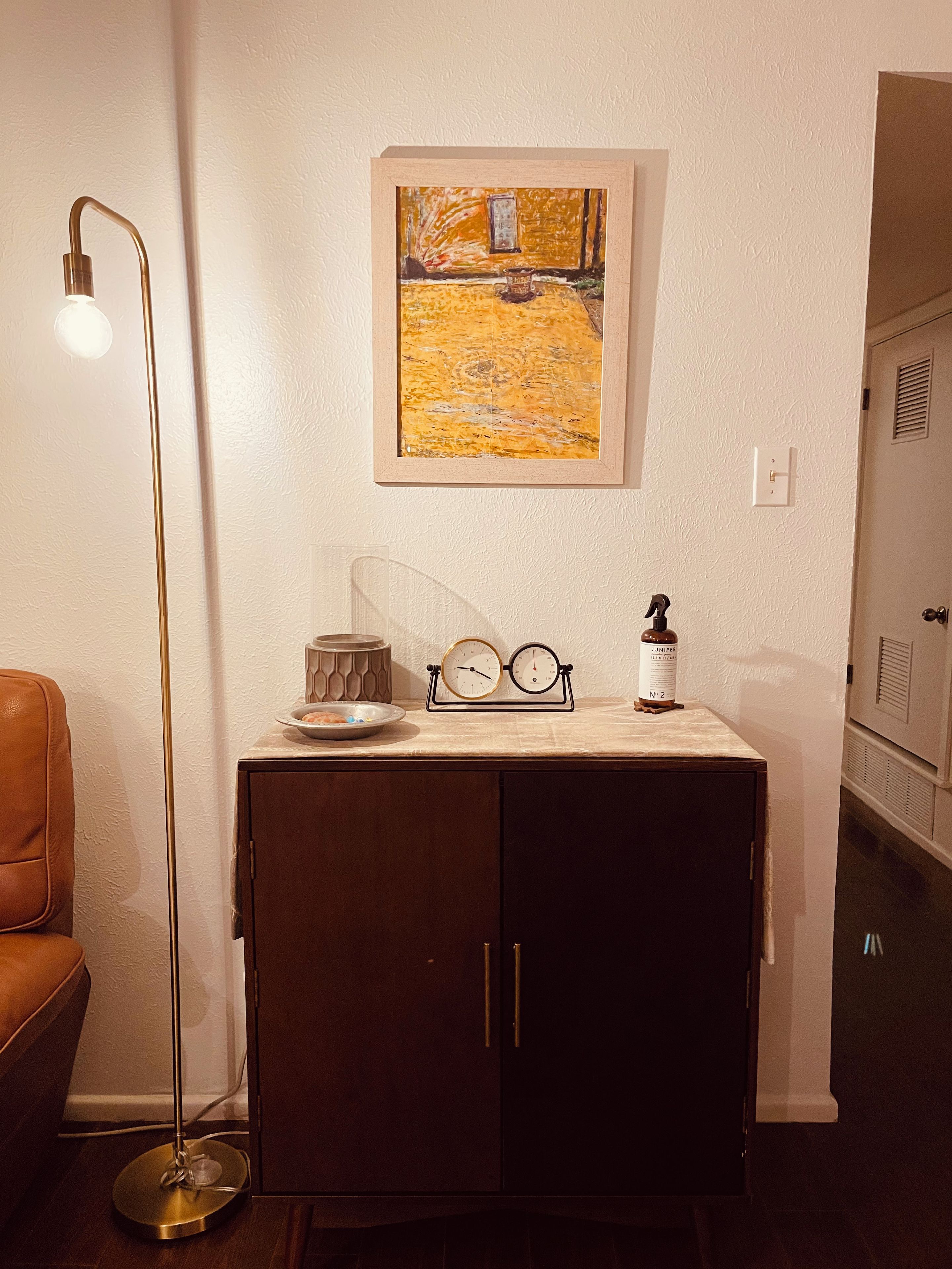 Een kamer met een donkere houten kast, een vloerlamp, een lijst met een schilderij, en een kleine tafel met een fles en glazen.