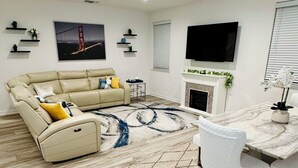 Una sala de estar moderna con un sofá seccional color beige, una chimenea, un comedor con una mesa y sillas blancas, y un televisor montado.