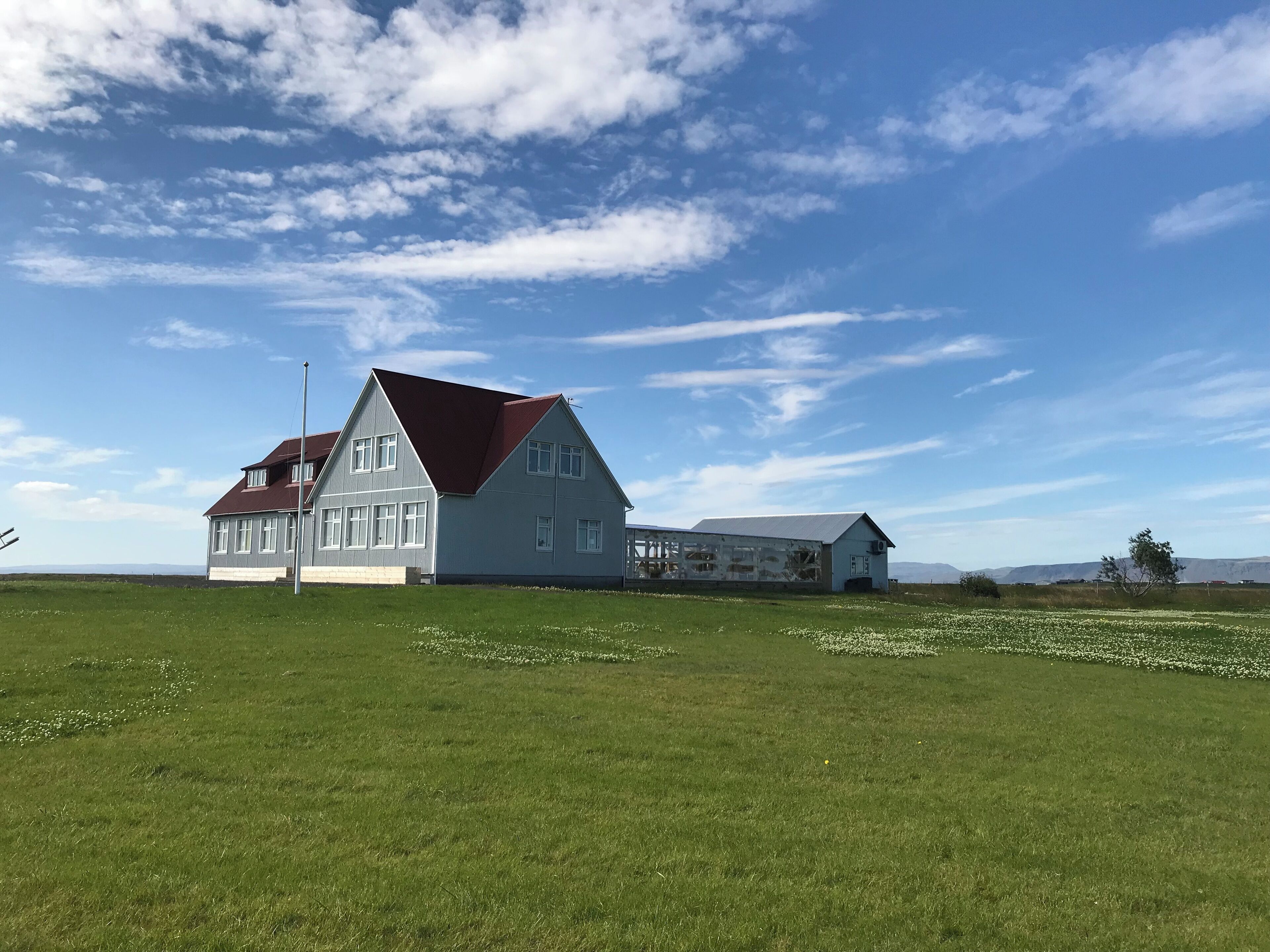 A large, two-story house with a red roof and white walls, situated in a grassy field with a clear sky.