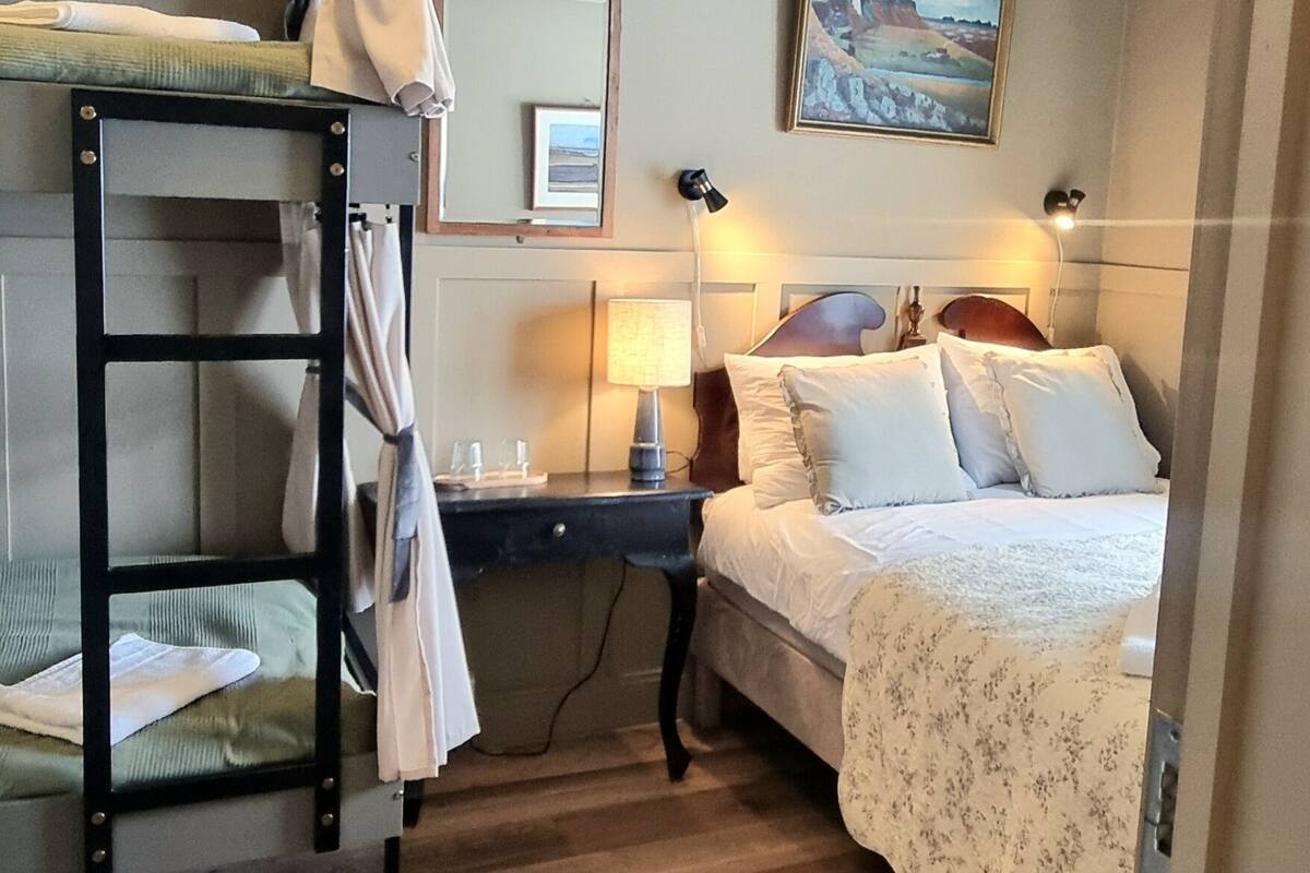 A bedroom with a bunk bed, a bedside table with a lamp, a wall-mounted mirror, and a painting.