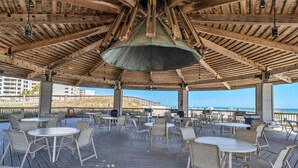 Een terras buiten met houten tafels en stoelen, een overdekt gedeelte met een metalen dak en uitzicht op de oceaan.