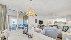 Een ruime woonruimte met een witte eettafel, een blauwe bank en een witte fauteuil. Er is een groot raam met gordijnen, een plafondventilator en een decoratieve hanglamp.